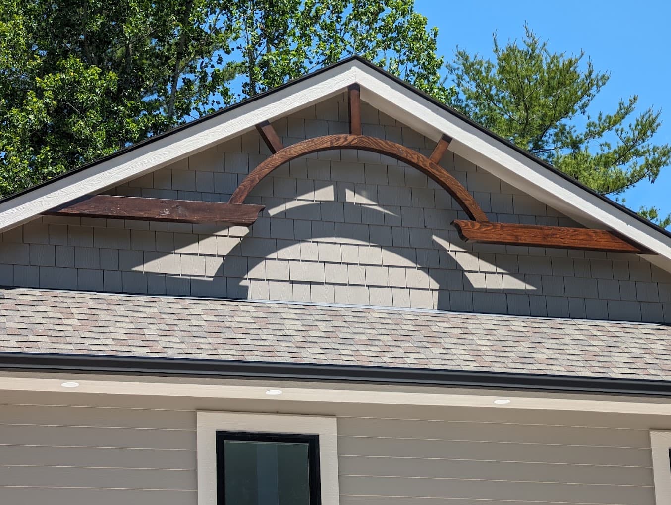 Finished stained wood arched bracket spanning a gable peak between two roof posts, set against cedar shake siding