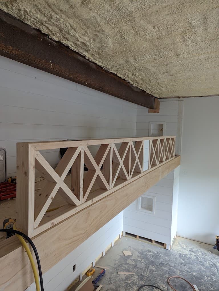 An interior loft railing under construction: square wood posts with X-pattern crossbuck panels between them, spray-foam insulation still exposed on the ceiling above, a drill and cord left on the subfloor.