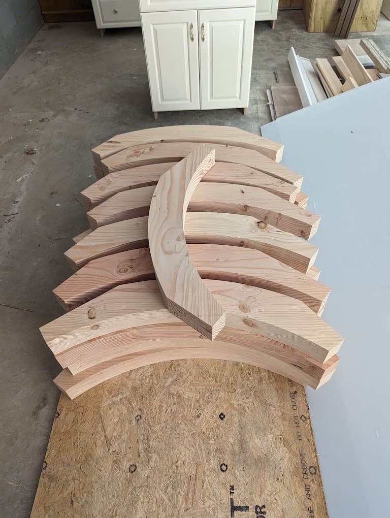 A batch of identical curved wood segments cut from dimensional lumber, stacked on a sheet of plywood in a shop, one segment turned to show its profile.