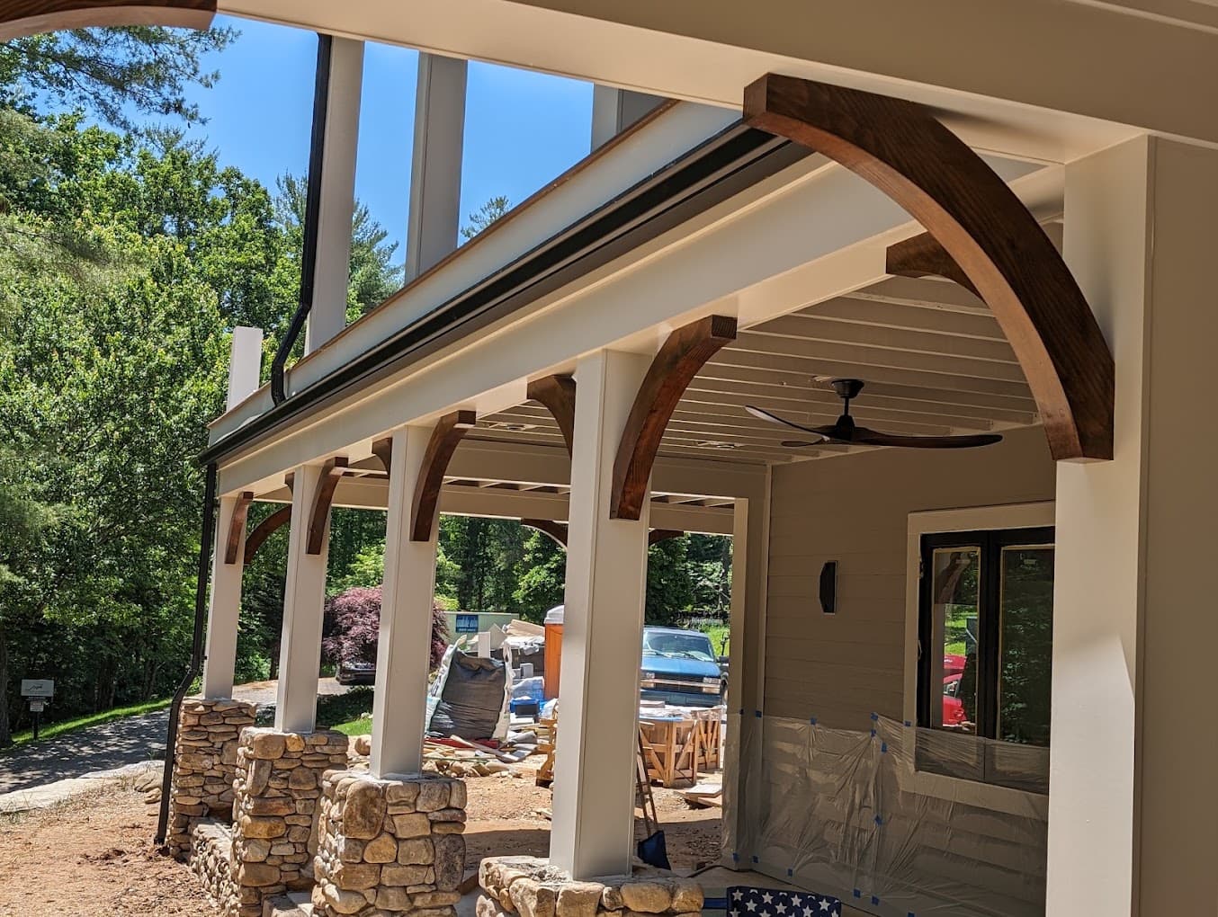 A row of curved wood knee braces running post to beam under a porch roof, stone-clad columns below, stained dark to match the porch ceiling, a ceiling fan overhead.