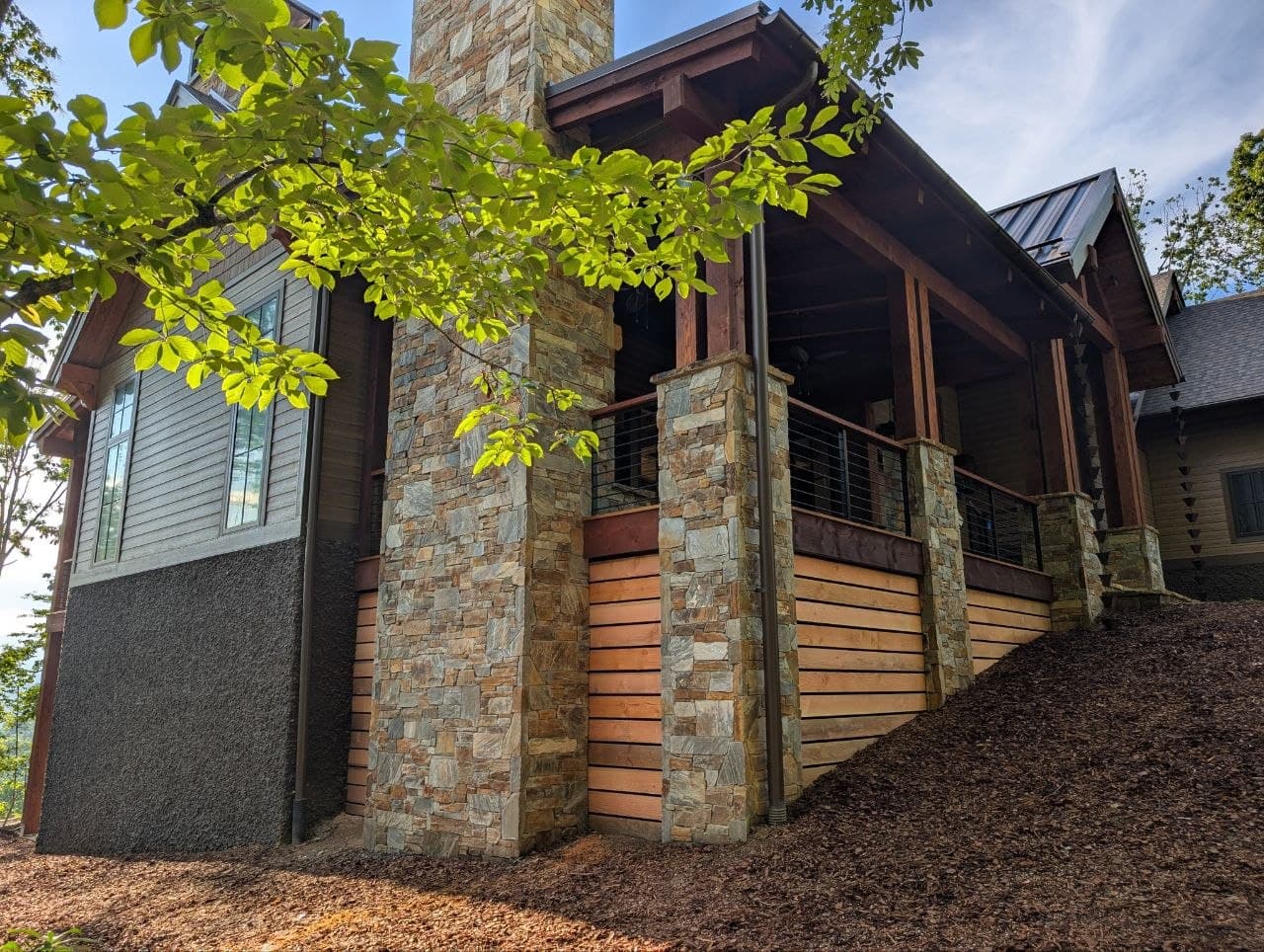 Horizontal skirting boards in raw, unstained wood beneath a deck, running past a tall stacked-stone chimney and stone porch columns with a cable-rail deck above.