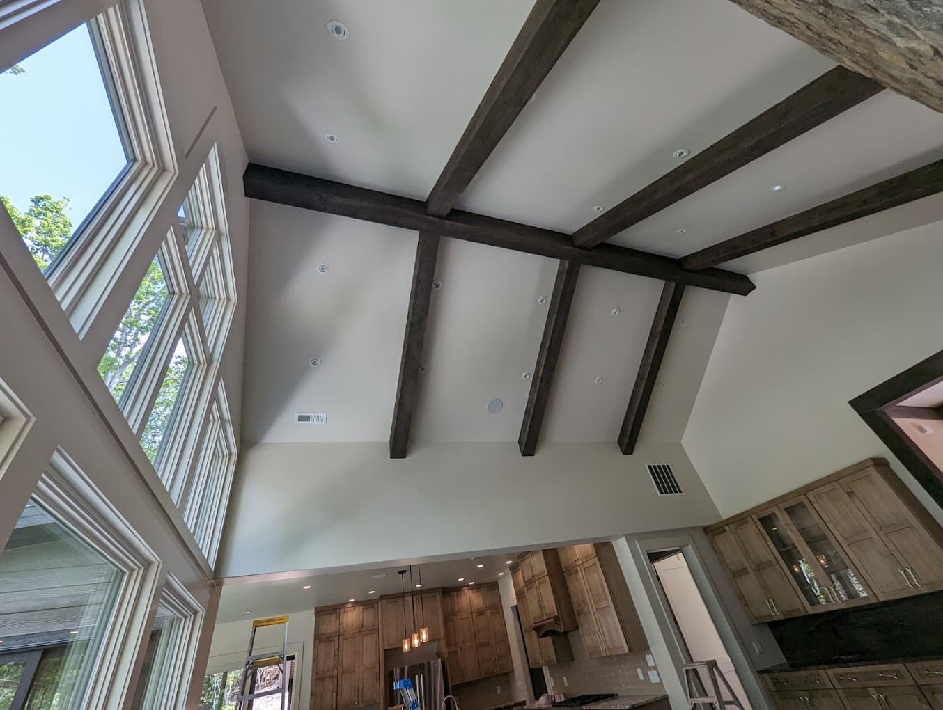 Dark stained wood beams crossing a vaulted white ceiling and meeting at a center ridge, recessed lights set in the panels between them, a kitchen visible below.